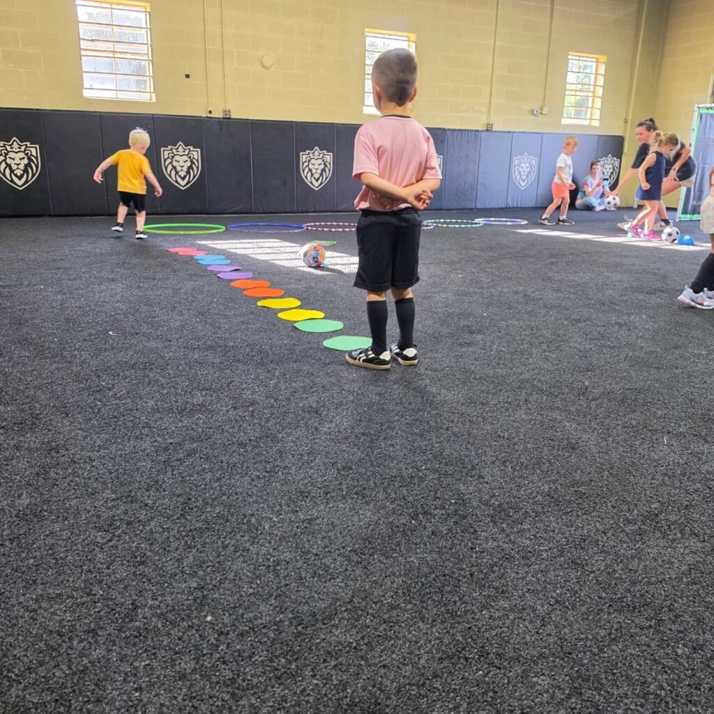 Child wearing pink shirt and black shorts stands on a line of colorful circular mats in a gym, with other kids and a coach in the background.